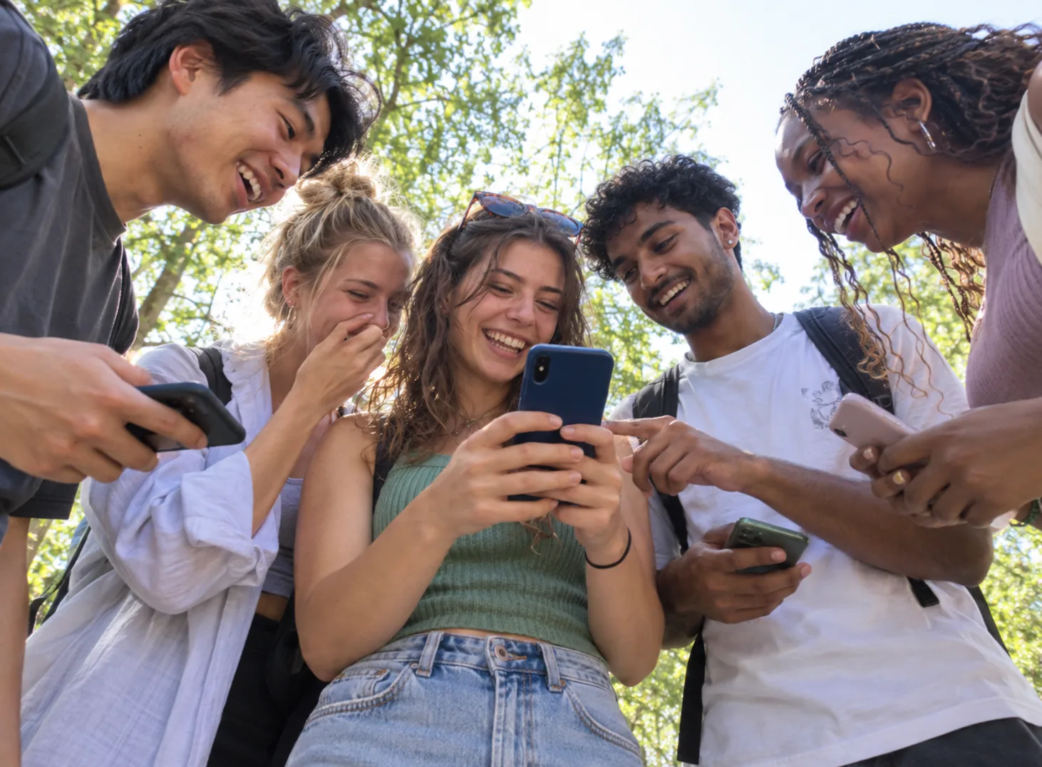Cinq jeunes rient en regardant un téléphone, sous les arbres